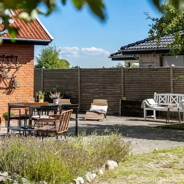 En terrasse med stole og et bord står ved et hus med røde mursten. Solskin oplyser området, der er omgivet af en træhegn og forskellige planter. Der ses flere huse i baggrunden.