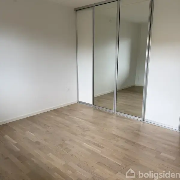 Tomt rum med trægulv, reflekteret i store spejldøre på en indbygget garderobe. Hvidmalede vægge uden dekoration, hvilket skaber et lyst og åbent udseende.