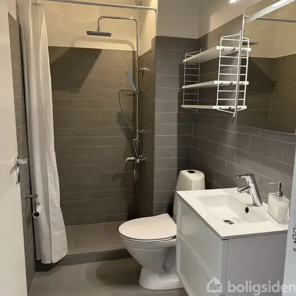 Et moderne badeværelse med brusekabine og grå fliser. Der er et hvidt håndvaskskab med en indbygget hane. Over håndvasken er et spejl og en hylde. En toiletpotte står ved siden af.