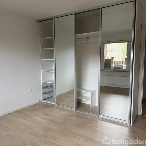 Skab med spejldøre står åbent i et værelse med trægulv, der viser hylder og en lille bænk. Et vindue lukker dagslys ind, og en radiator ses under vinduet.