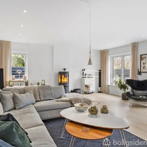 En lys stue med stor, grå hjørnesofa fyldt med puder. Sofaen står ved et ovalt sofabord. I hjørnet står en brændeovn. Vinduer lukker dagslys ind, og planter pynter rummet.