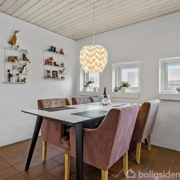 Spisebord med fire lyserøde stole står i en lys spisestue. En designerlampe hænger over bordet. Små hylder med dekorative figurer pynter væggen. To vinduer med planter bagved.