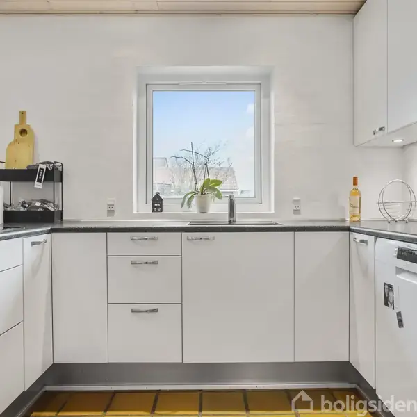 Hvidt køkken med hvide skabe, hvor en opvaskemaskine og ovn er indbygget. En plante står i vinduet over vasken. Bordpladerne er sorte, og gulvet er gult flisebelagt.