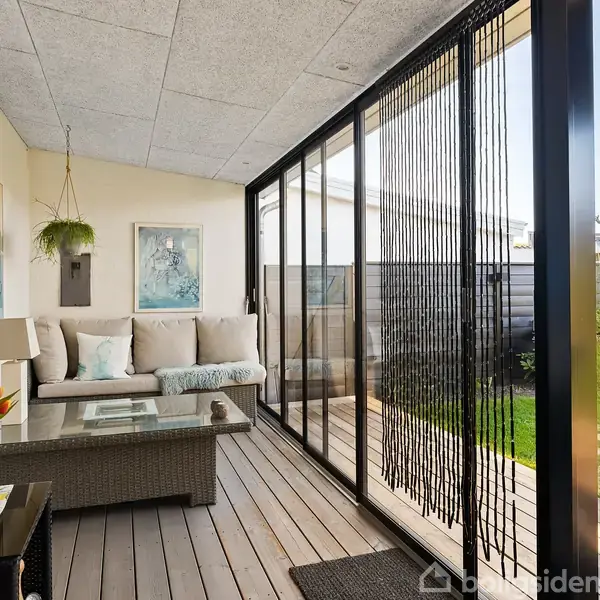 Sofa med puder står på en overdækket terrasse. Et glasbord foran sofaen. Store glaspartier og hængende plante skaber en lys og hyggelig atmosfære med udsigt til haven.