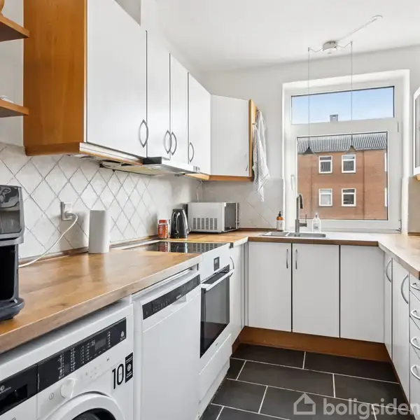 Køkken med træbordplader, hvide skabe, sort og hvid mosaikvæg. Vaskemaskine og mikrobølgeovn synlige. Ud af vinduet ses en rød murstensbygning. Bøger på hylde øverst.