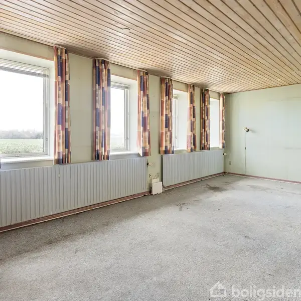 Et tomt rum med et træloft har fire vinduer med farvestrålende gardiner langs én væg. Væggene er lysegrønne, og der er en radiator nedenunder vinduerne. Ingen tekst er til stede.