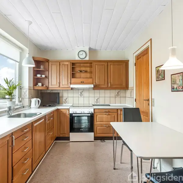 Køkken med træskabe og hvidevarer, inklusive en ovn. Et spisebord med stole står ved siden af. Vindue med planter til venstre; vægur og malerier på væggen.