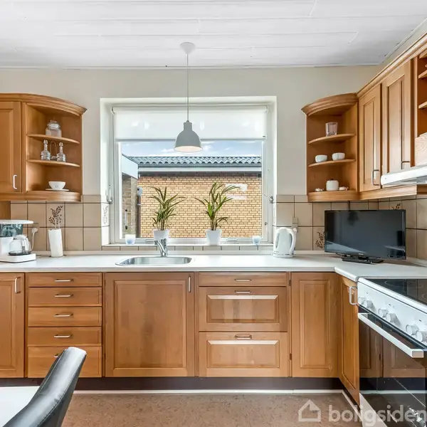 Køkken med træskabe, kaffemaskine og komfur. Står stille med en stor vinduesåbning mod en murstensbygning udenfor. Lyse elementer og dekorationer på hylderne. Ingen tekst fundet.