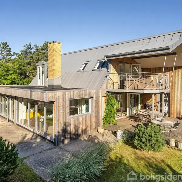 Et moderne hus med fladt tag og store vinduer står i en velholdt have. Solen skinner, og der er træer og buske i baggrunden.
