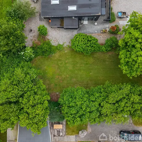 Græsareal er omgivet af tætte grønne træer og buske. Ovenfor ses et hus med mørkt tag omgivet af en grusbelagt haveplads.