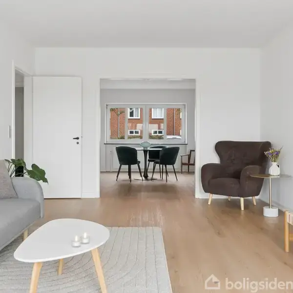 En grå sofa og et hvidt sofabord står i en lys stue. I baggrunden ses et spisebord med stole foran et vindue. En brun lænestol står til højre.