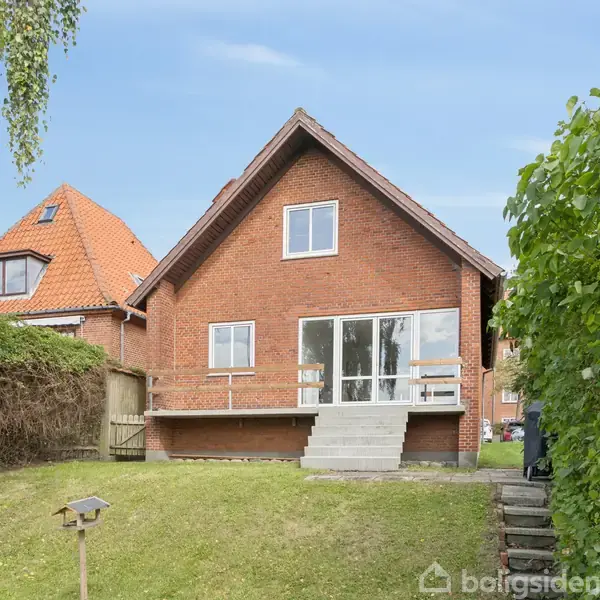 Rødt murstenshus med skråtag ligger i en grøn have med hæk og trappe. Ved huset er en terrasse med glasdør og trapper. Solrig dag med en blå himmel.