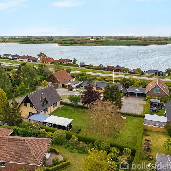 Flere villaer ligger i et grønt boligområde nær en bred flod. Haverne er velplejede, og der er en vej langs flodbredden.ğ