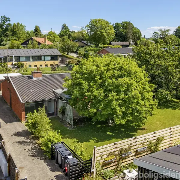 En murstensvilla med fladt tag står i en frodig have med græsplæne, store træer og en indkørsel med bil. Omgivet af hegn og andre huse i et grønt boligområde.