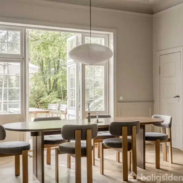 En spisestue med et langt træbord og seks polstrede stole. Store glasdøre åbner ud mod en terrasse med udsigt til en frodig have. Enkel lampe hænger over bordet.