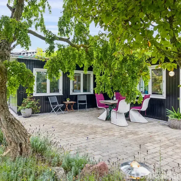 Et stort, frodigt træ kaster skygge over en hyggelig terrasse med bord og lyserøde stole. I baggrunden ses et sortmalet hus med hvide vinduesrammer omgivet af grøn beplantning.