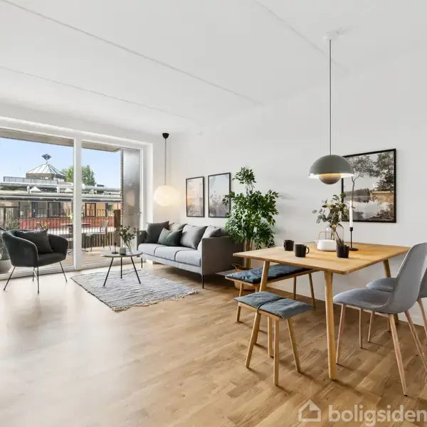 Et moderne, lyst køkken-alrum med trægulve strækker sig mod en stue med sofa, stuebord og planter. Store vinduer åbner mod en balkon og udsigt til byen.