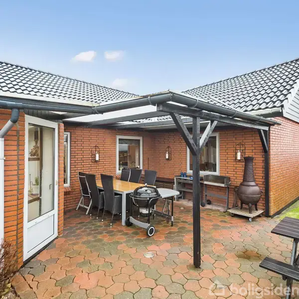 En udendørs terrasse med et sort grillbord og en spisebordssæt under en overdækning. Omgivet af en rød murstensbygning og en grøn græsplæne.