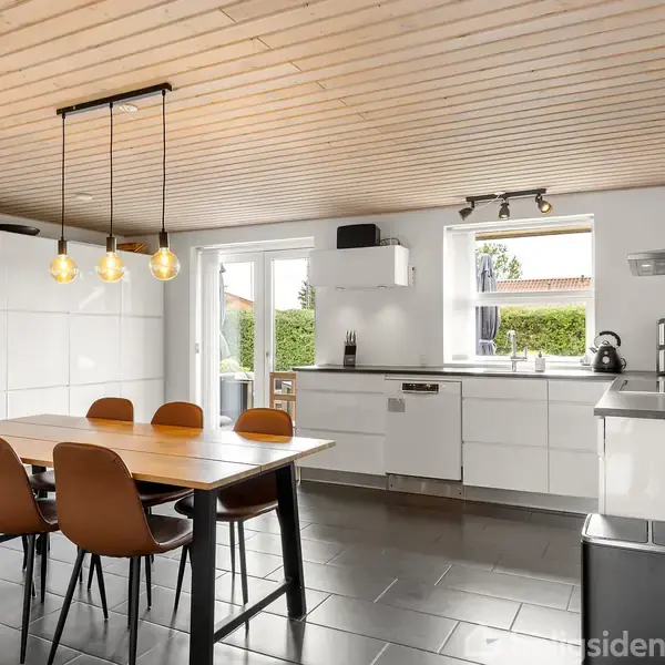 Et moderne køkken med hvide skabe og apparater. Et træbord med seks stole står i midten. Store vinduer og en glasdør giver udsigt til en terrasse.