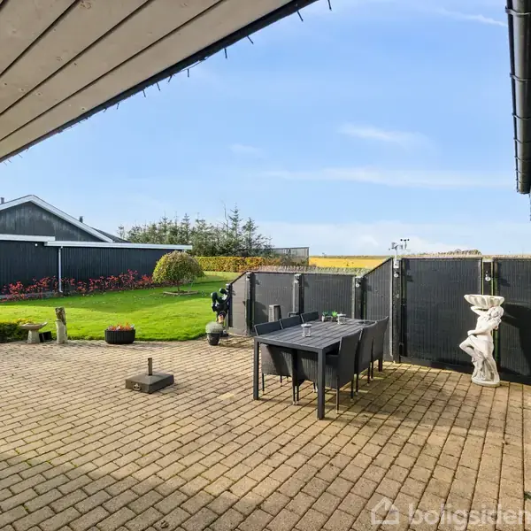 En terrasse har et sort bord med seks stole placeret på fliser. I baggrunden er en grøn græsplæne med buske og et mørkt hus. Skyfri himmel.