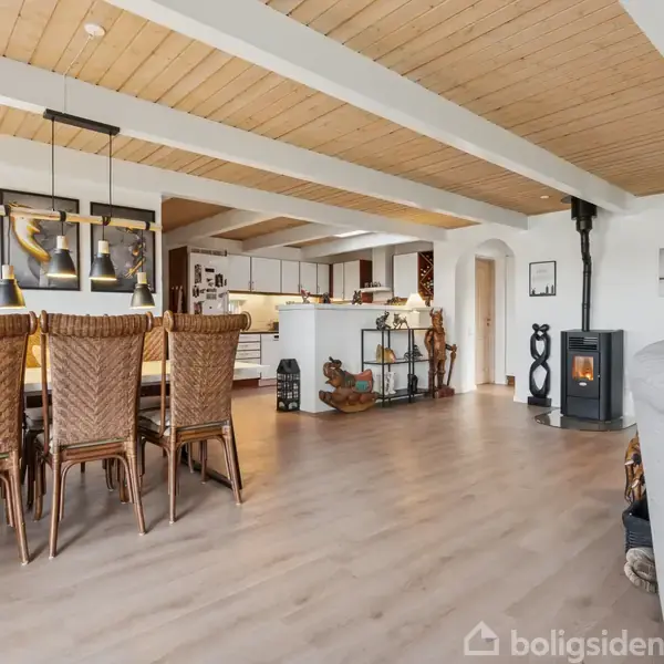 En stor stue med spisebord og seks fletstole står foran et åbent køkken. Væggene har kunst, og et brændefyr brænder i hjørnet, hvilket skaber en hyggelig atmosfære.