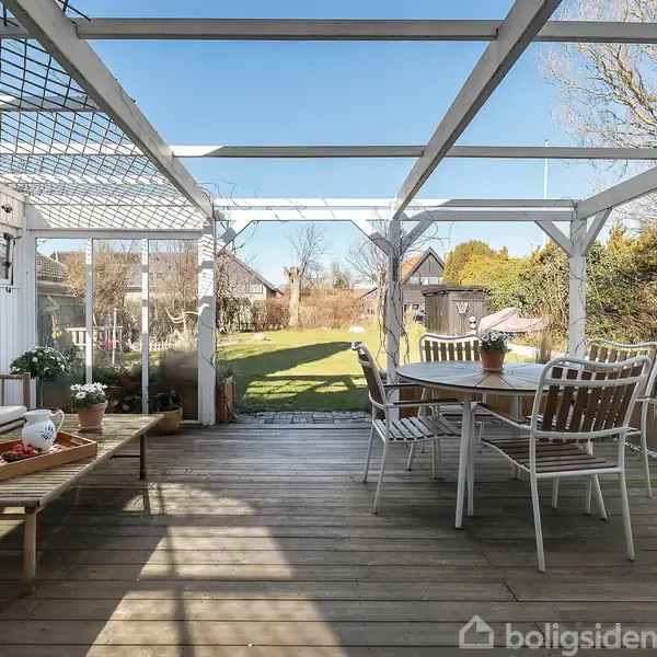 Terrasseområde med træmøbler, inklusive en rund spisebordssæt. En bænk står nær et hus, der er delvist skygget af pergolaoverdækning. Baggrunden er en have med græsplæne og træer.