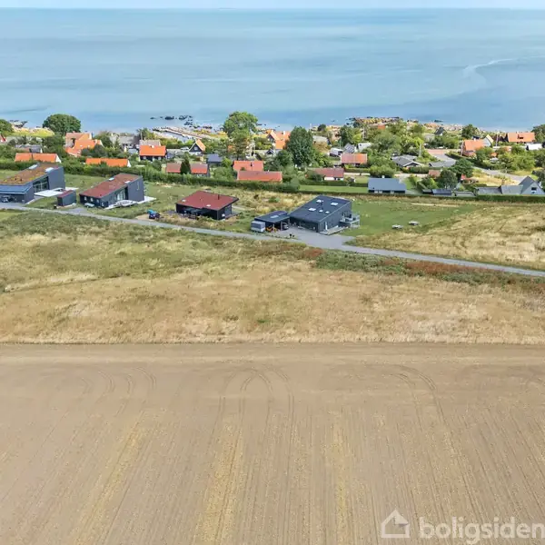 Landsbyhuse står stille i en bakket, landlig kystnær panorama, omgivet af gyldne marker og havudsigt i baggrunden, med spredte grønne træer ved horisonten.