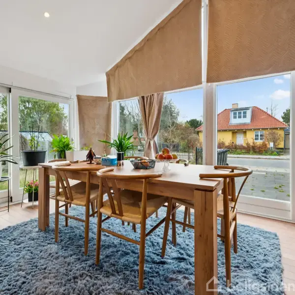 Et spisebord med seks stole står på et blåt tæppe i et solrigt rum med store vinduer og planter. Udenfor ses et gult hus og blå himmel.