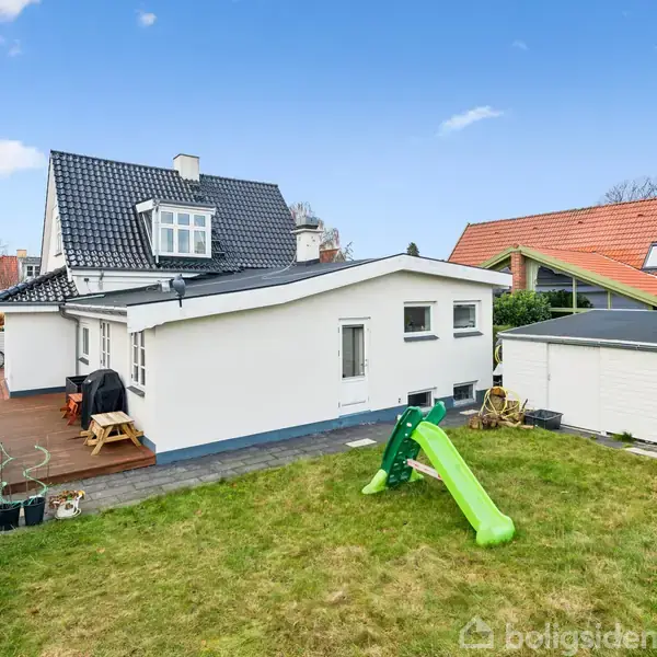 Huset står stille i en have med græs og en grøn rutsjebane. Ved siden af er der en terrasse med borde, stole og blomsterpotter under en klar blå himmel.