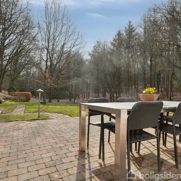 Et rødt hus med mørkt tag står stille i en snedækket have omkranset af træer. En sti fører op til huset, omgivet af buske og træer med bare grene.