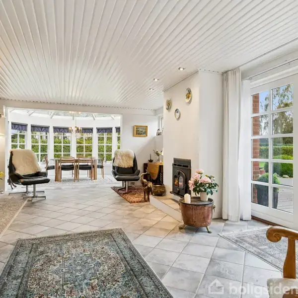 Stue med gulvtæppe og pejs, kører op ad loftet i et lyst rum. Store glasdøre og vinduer åbner mod en terrasse og have med grønne træer i baggrunden.