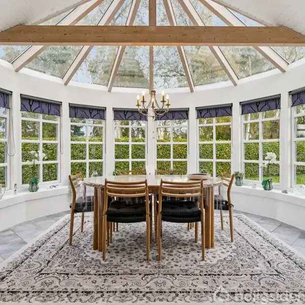 Et træbord med stole står central i et lyst rum med glasvægge og synligt loft. En lysekrone hænger over bordet, og rummet er omgivet af grønne buske udenfor.