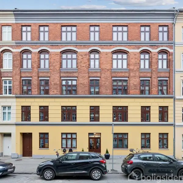 En etageejendom i en by med tre forskellige farver og arkitektoniske detaljer. Biler parkeret langs fortovet understreger ejendommens urbane miljø.