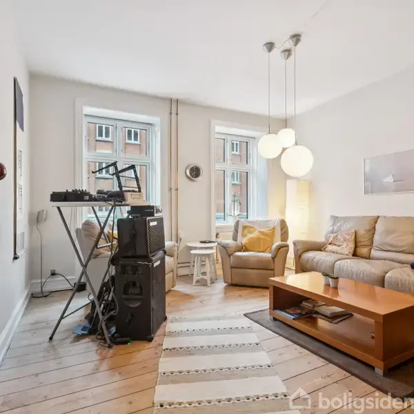 Stue med to store vinduer, hvor en beige sofa og sofabord står mod væggen. Et musikanlæg og keyboard står til venstre. Loftslampe hænger over sofabordet. Lys træparketgulv.