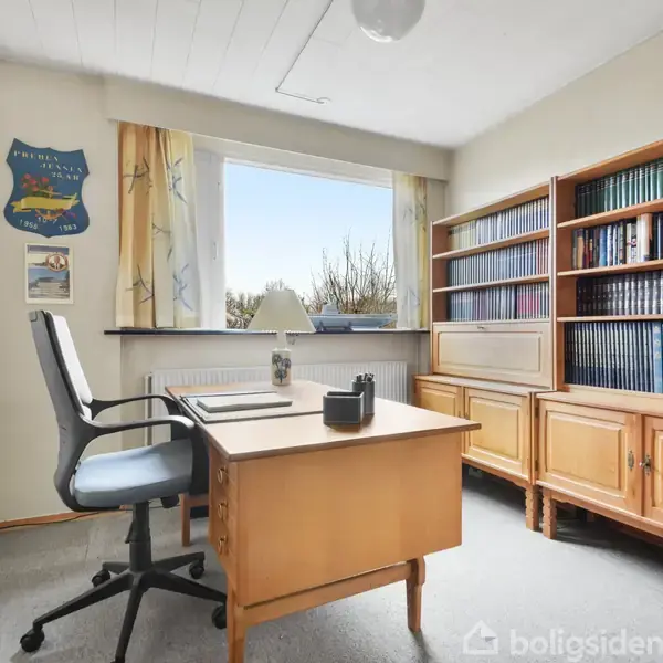 En kontorstol står ved et skrivebord med en lampe og notesblok, omgivet af bøger på en reol. Værelsesvinduet er åbent og lader naturligt lys komme ind.