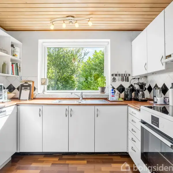 Et lyst køkken med hvide skabe og træbordplader. En ovn og opvaskemaskine er indbygget. Et stort vindue viser en grøn udsigt, omgivet af diverse køkkengrej og apparater.