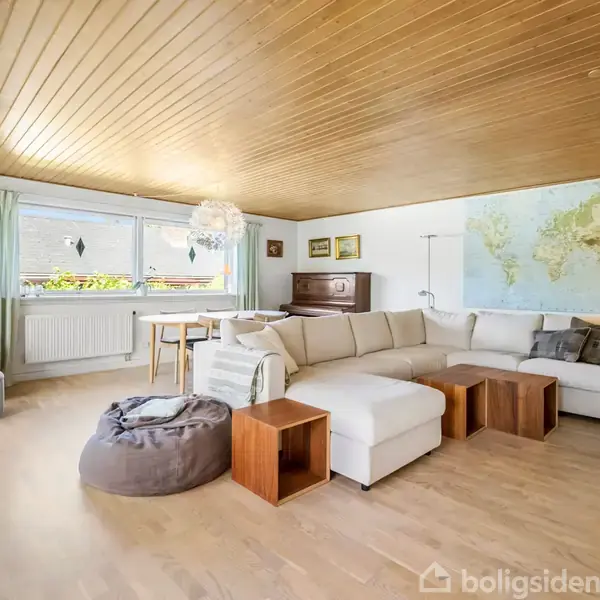 Stor stue med hvid hjørnesofa og en grå sækkestol foran et kort på væggen. En spisestue er bagved med et klaver til venstre og stort vindue med grønne gardiner.