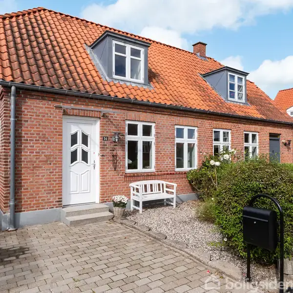 Et rødt murstenshus med hvide vinduer og orange tegltag står foran en flisebelagt indgang. En hvid bænk og grønne buske pryder forhaven, som omgives af en sort postkasse.