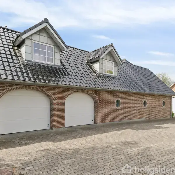 En murstensbygning med mørkt tegltag har to garageporte og to loftsrumsvinduer. Bygningen står på en brolagt indkørsel i et boligområde med andre huse omkring.