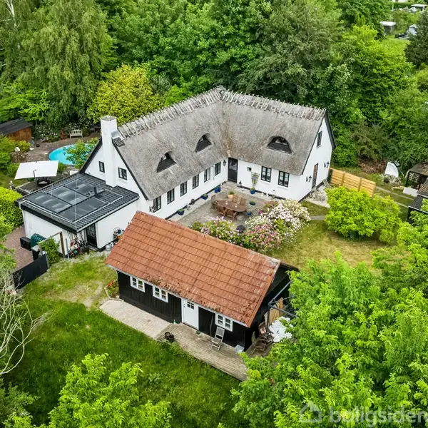 Stråtækt hus og udhus omgivet af frodig have. Huset har en indre gårdhave med blomsterbede. I baggrunden ses svømmebassin og tæt skovbevoksning.