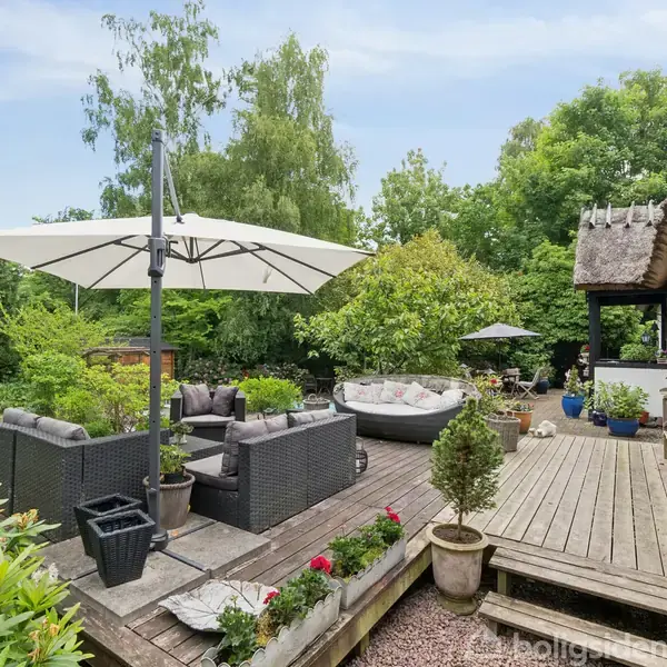 Terrassemøbler er placeret under en stor parasol. Omkring er der frodig beplantning og træer. En træterrasse strækker sig langs et hus med stråtag, hvor flere planter står i krukker.