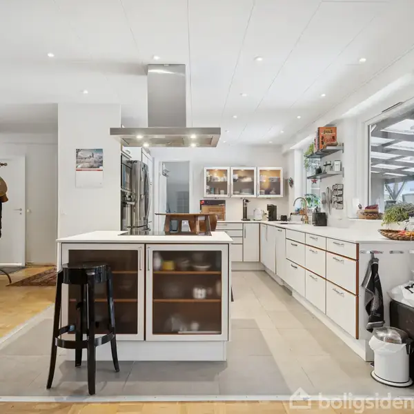 Et moderne køkken med en central kogeø og sort barstol. Omkring er hvide skabe og hylder fyldt med køkkengrej og dekorationer. Lyse parketgulve og store vinduer skaber en lys atmosfære.