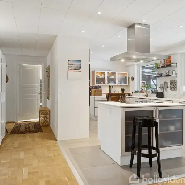 Et moderne køkken med hvidt interiør og en central ø, der har en sort skammel foran. Rummet er lyst med store vinduer og udsigt til en overdækket terrasse.