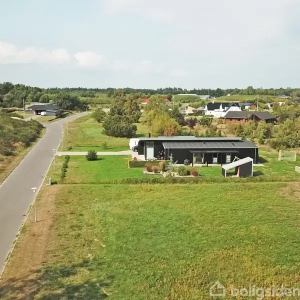 En moderne, sort rektangulær bygning står på en velplejet græsplæne, omgivet af et fladt landskab og en stille vej. Flere huse ligger i det fjerne.