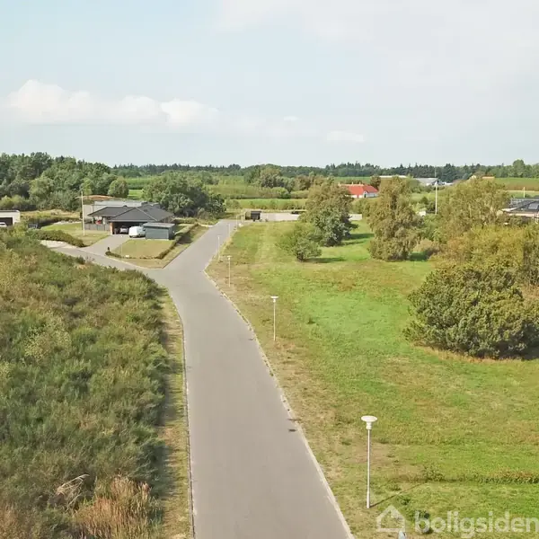 En stille vej strækker sig gennem et grønt boligområde med enkelte huse og træer, omgivet af åbne marker og skove i horisonten under en blå himmel.