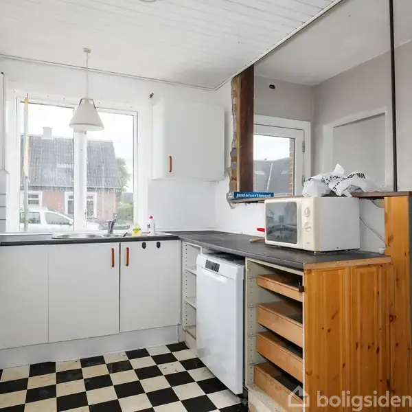Køkken i et lyst rum med sort-hvidternede fliser. Et køkkenbord med hvidt skab, opvaskemaskine og mikrobølgeovn. I baggrunden ses en trappe og store vinduer mod husfacader.