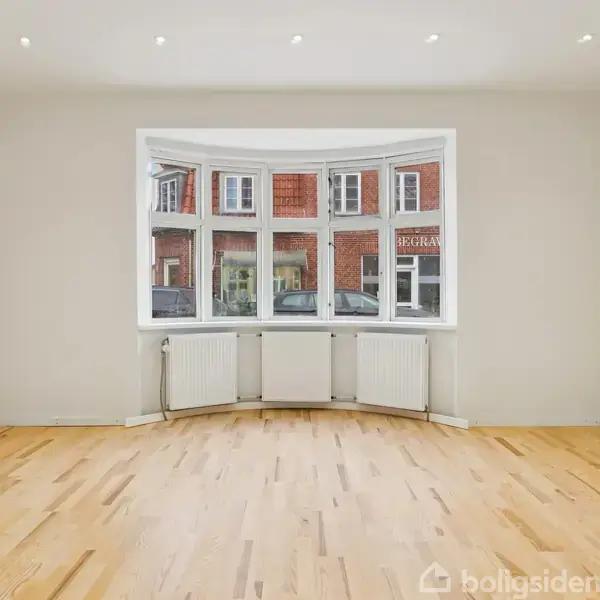 Et tomt rum har et stort, buet vindue, fire radiatorer under vinduet og lyst trægulv. Udenfor ses en rød murstensbygning med tekst “KORAN” synlig i vinduerne.