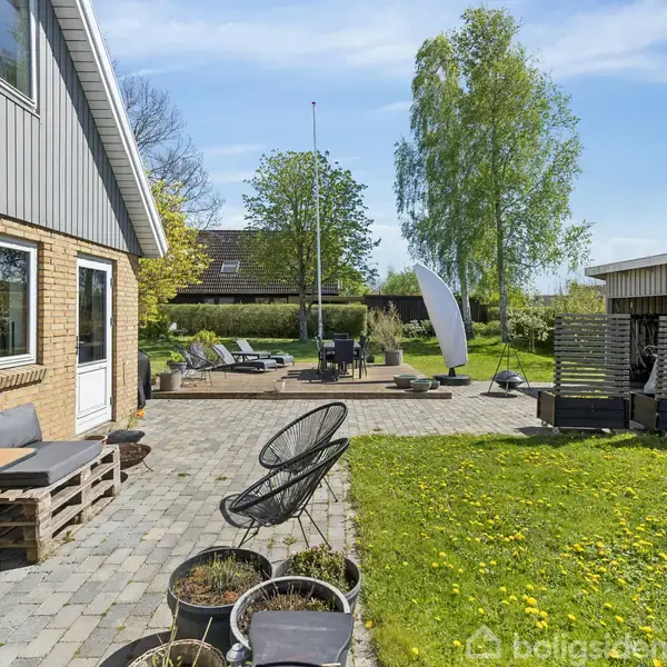 Terrasse med træmøbler og puder står langs husmur. Rundt om er der en have med grøn græsplæne og høje træer i baggrunden. Fliser dækker resten af terrassen.