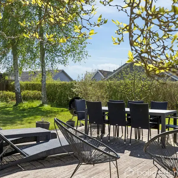 Terrassemøbler står arrangeret på en træveranda, omgivet af grønne træer og en trimmet græsplæne. I baggrunden ses en række huse, der tilføjer til en fredfyldt, solrig havedag.