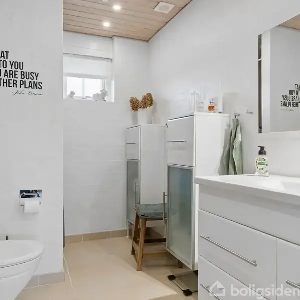 Toilet i hvidt badeværelse med vask og skab. Citat på væggen: "Life is what happens to you while you are busy making other plans." Vindue med lysindfald, håndklæder på stang, lyse trægulve.